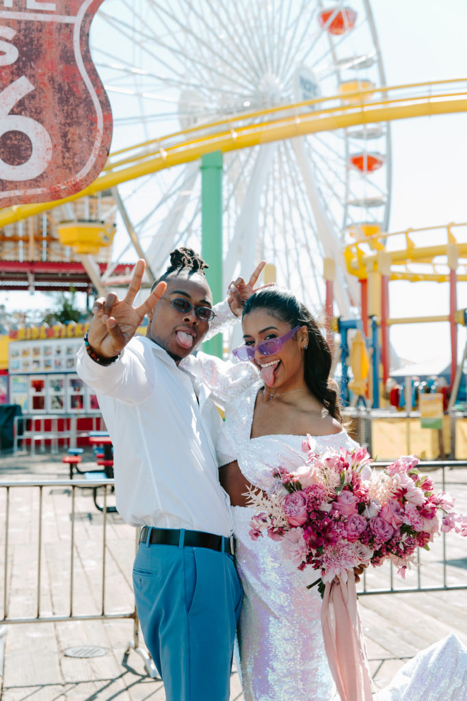 Newly wed couple standing among the fair rides at Santa Monica Pier sticking their tongues out and throwing up peace signs during their urban Los Angeles elopement