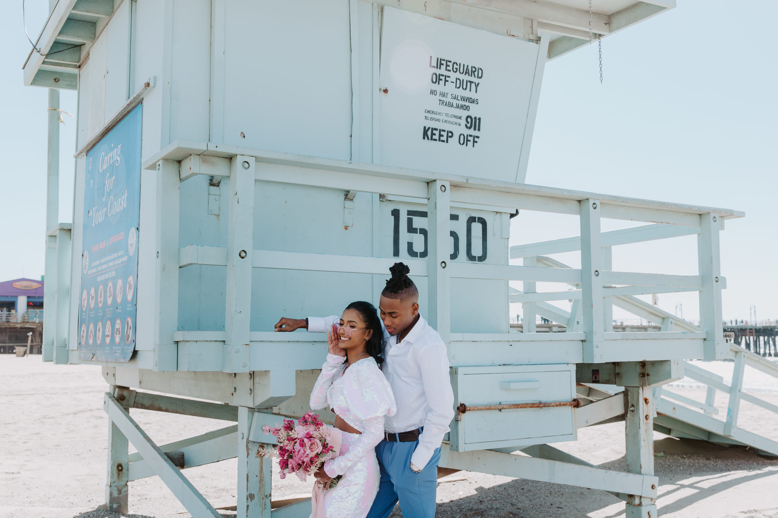 A newlywed couple leaning up against a lifeguard stand the bride is in a pink two piece dress holding a bouquet of pink flowers and the man standing behind her is in bright blue dress pants and a white shirt during their Santa Monica pier wedding in California
