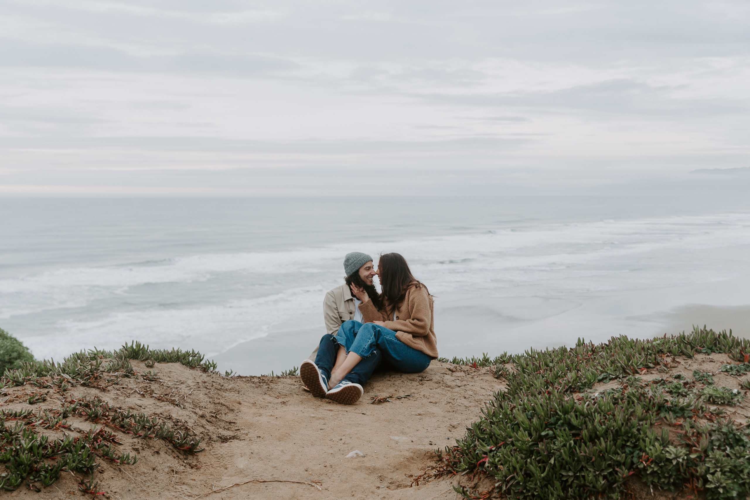 Newly engaged couple sitting on a cliff overlooking the ocean while the woman is holding gently on to her partners cheek during their San Francisco engagement photos