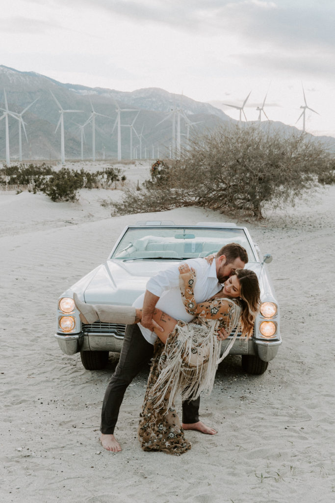 Man dipping his partner in front of a vintage car as they are surrounded by windmills in the Palm Springs desert during their California engagement photos