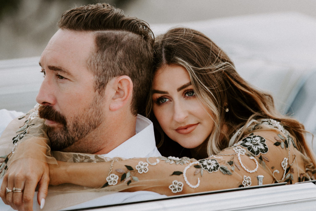 A guy sitting in the front seat of a convertible with his partner sitting in the back but she's leaning in with her arms wrapped around him during their edgy engagement photos near Joshua tree in Palm Springs California