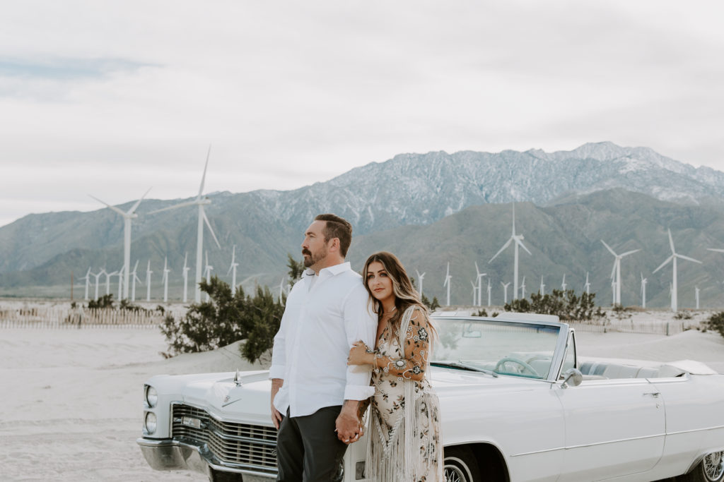 A couple standing in front of a vintage white Corvette convertible with a palm springs windmills in the far background during their engagement photos in the desert of southern California