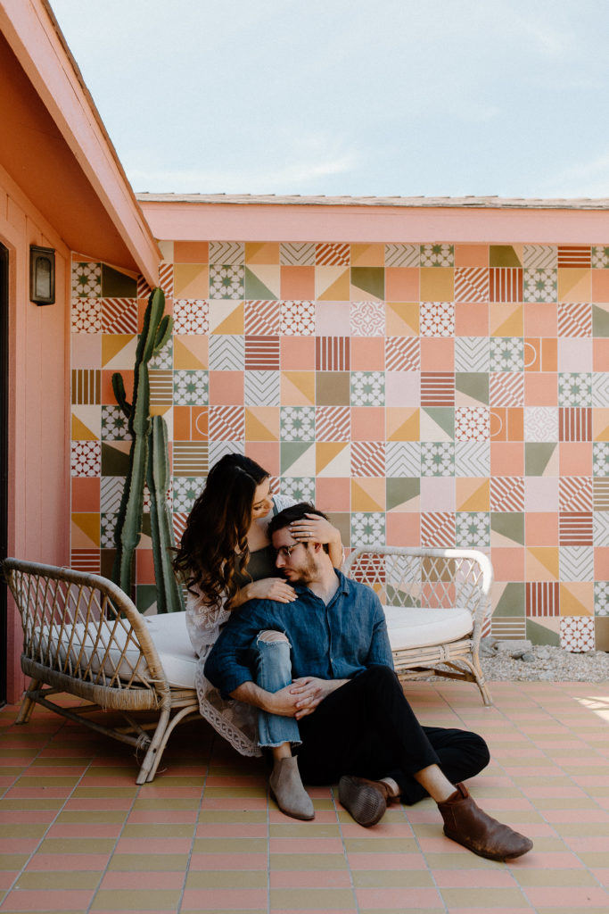A man and a woman sitting on their outdoor seating as the man is on the ground sitting between his partners legs and she is brushing her fingers through his hair during their Palm Springs couple session