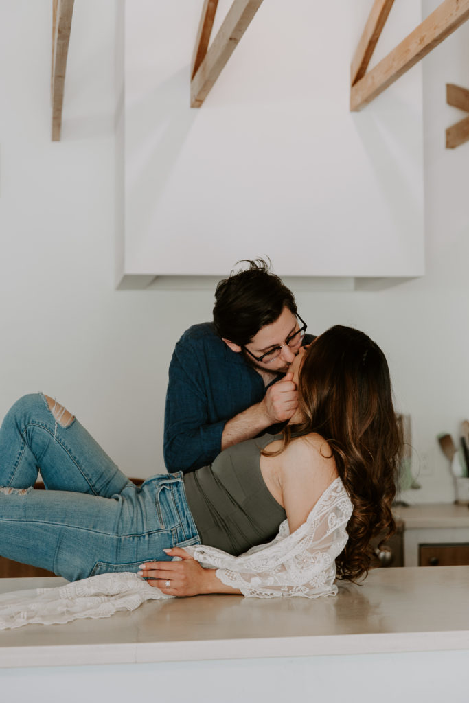 A woman laying on her kitchen counter with her partner standing over her and bringing her in for a kiss during their California couple session