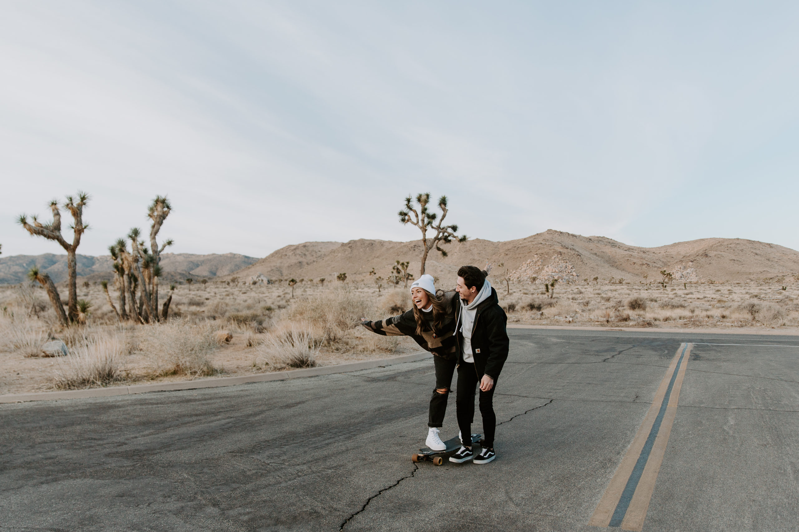 Man pushing his partner on a skateboard as she has her arms thrown wide with a big smile on her face during their adventurous engagement photos in California
