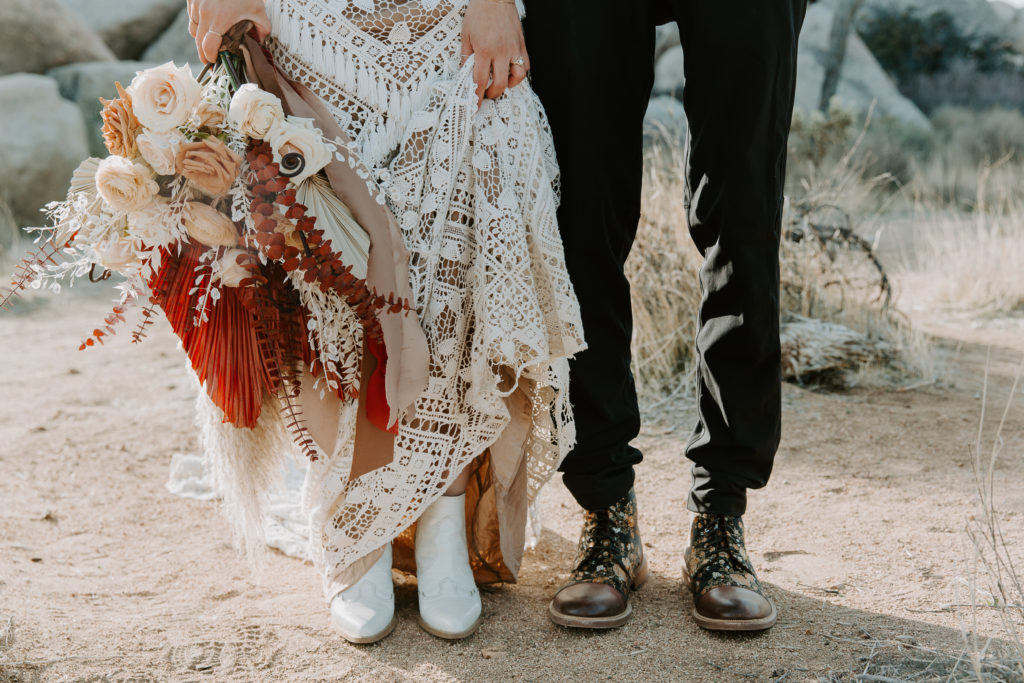 A couple standing side-by-side and wedding attire as the woman is holding up her dress to show off her white wedding boots during this couples Joshua tree destination wedding