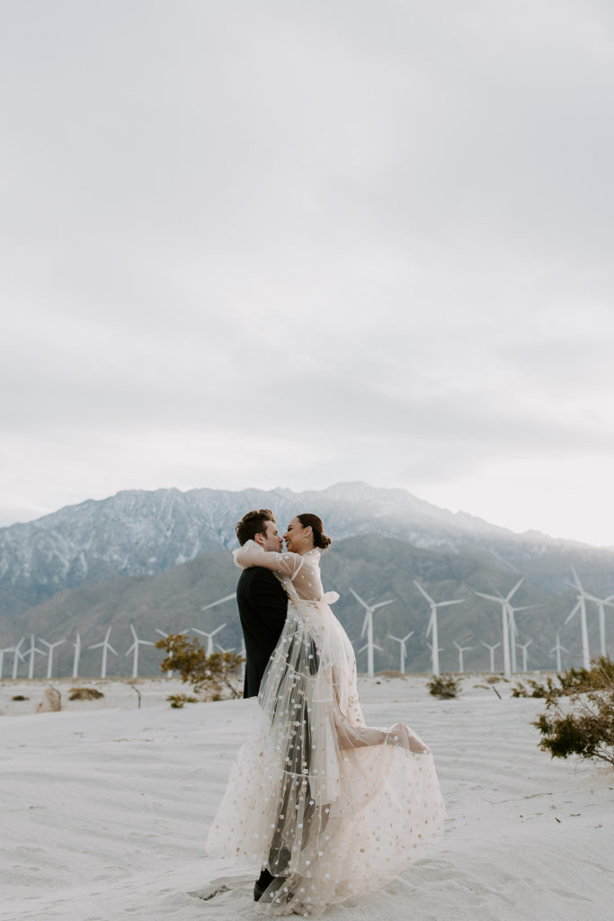 Man holding up his partner as she leans in to give him a kiss with the mountains and windmills in the background during their California elopement