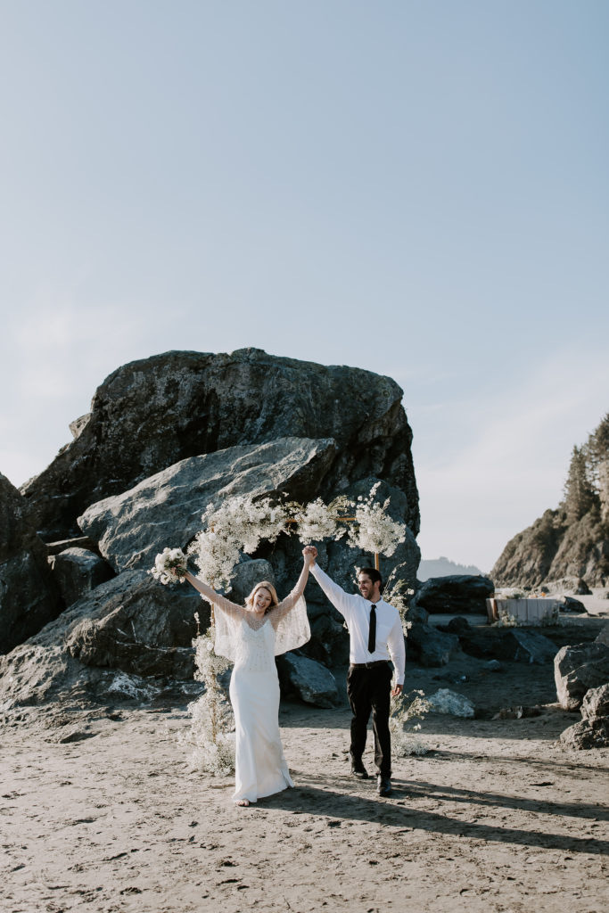 A couple celebrating in front of their beach arch after getting married during their california micro wedding