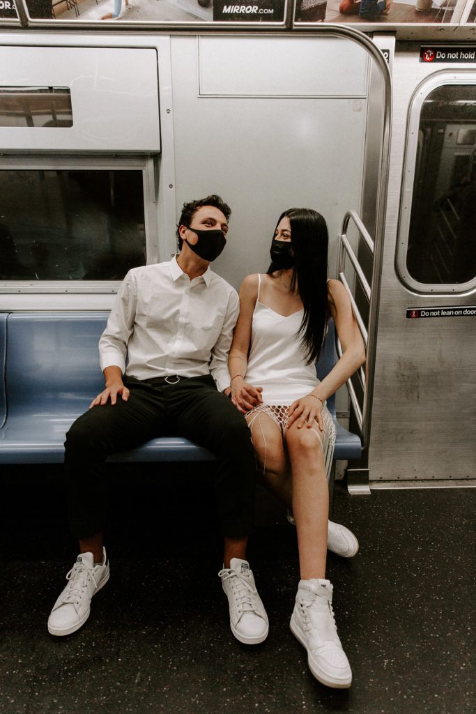 A man and a woman sitting on the subway holding hands as the woman is looking at her partner and her partner is looking at the signs during their NYC wedding