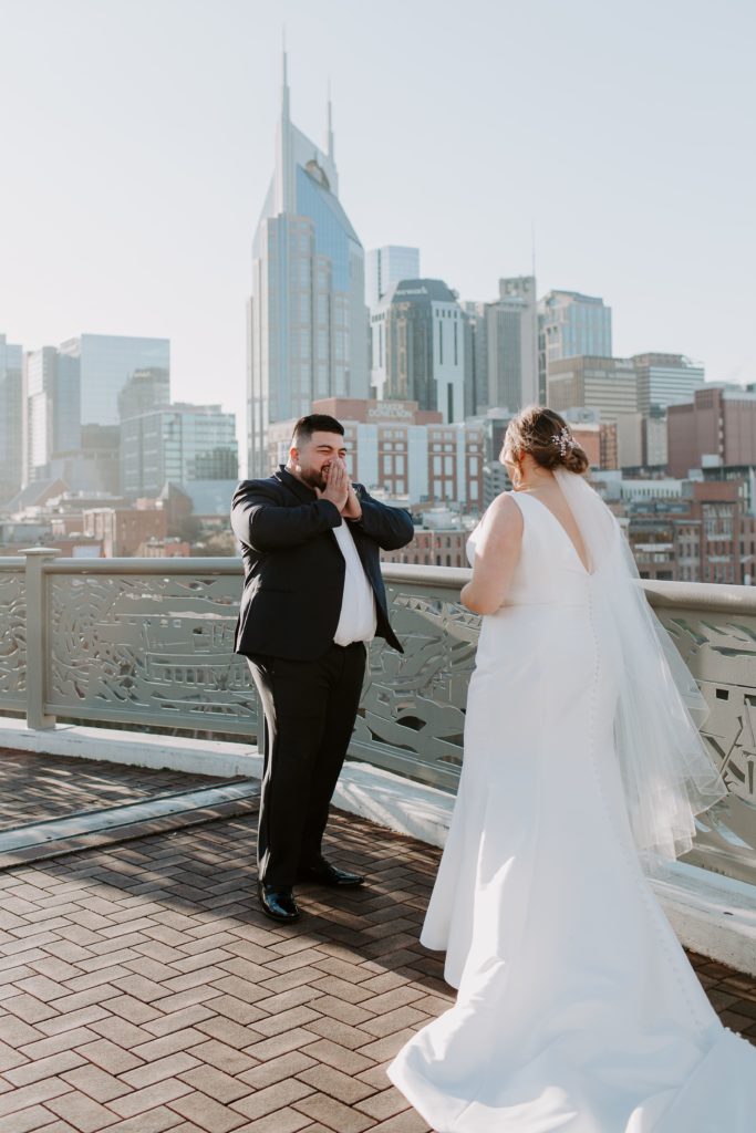 Man with his hands covering his mouth in awe of his bride after seeing her for the first time with the Nashville skyline in the background