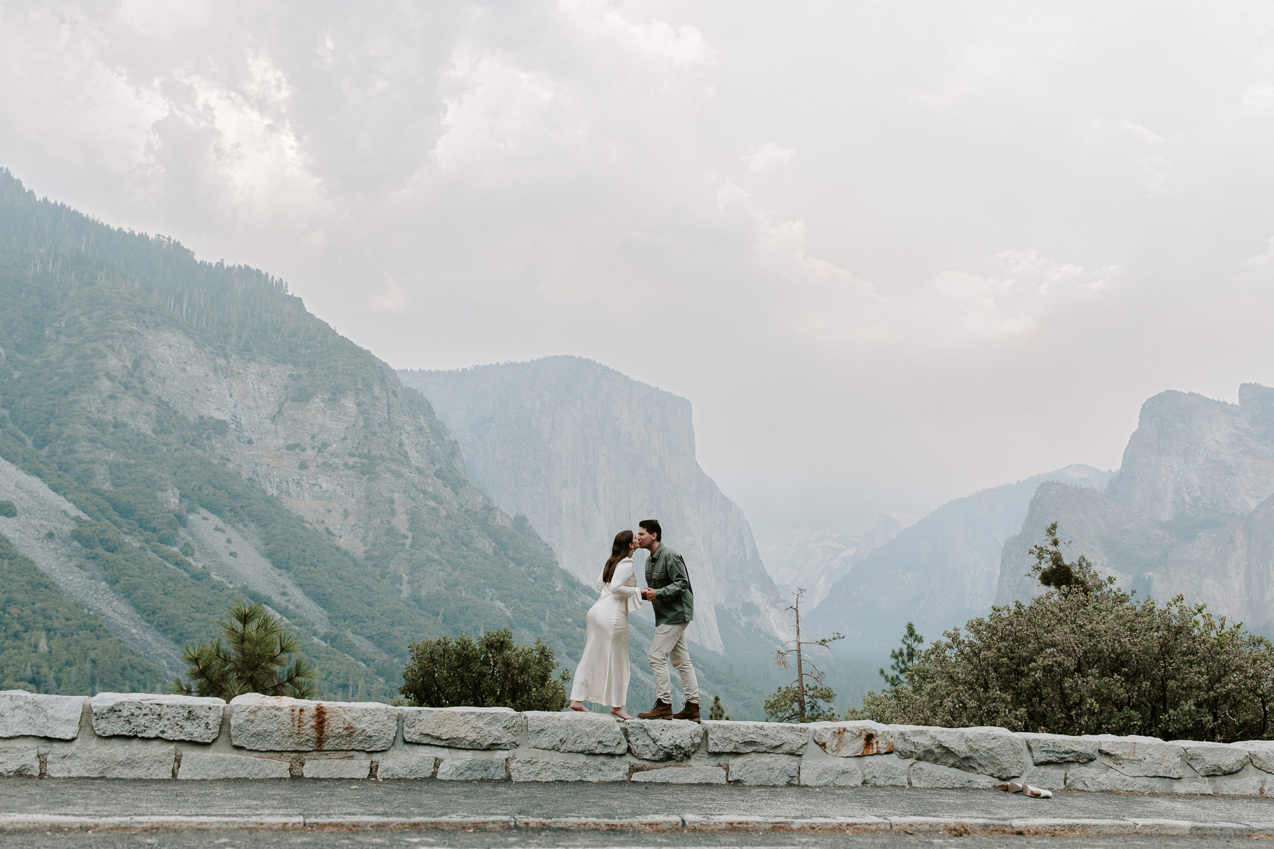 Couple standing on a wall sharing a kiss with Yosemite Valley in the background during their couple photos at the Tunnel View of Yosemite