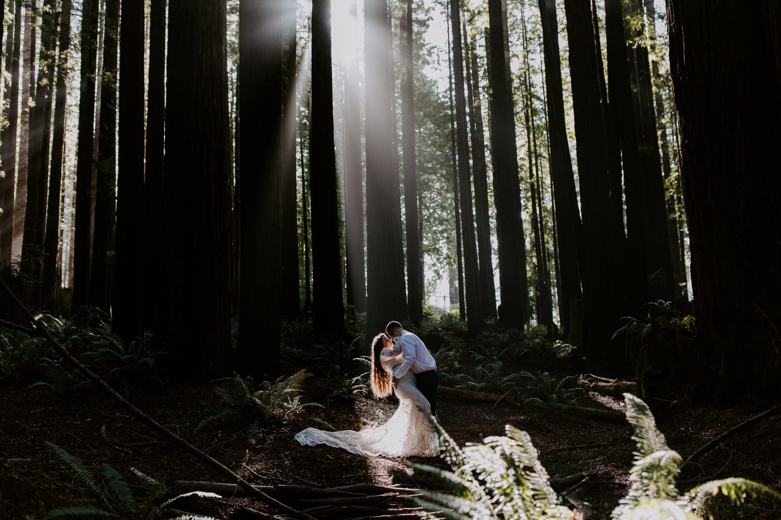 Groom dipping his partner for a kiss as the sun filters through the redwoods during their Northern California elopement in the Redwoods