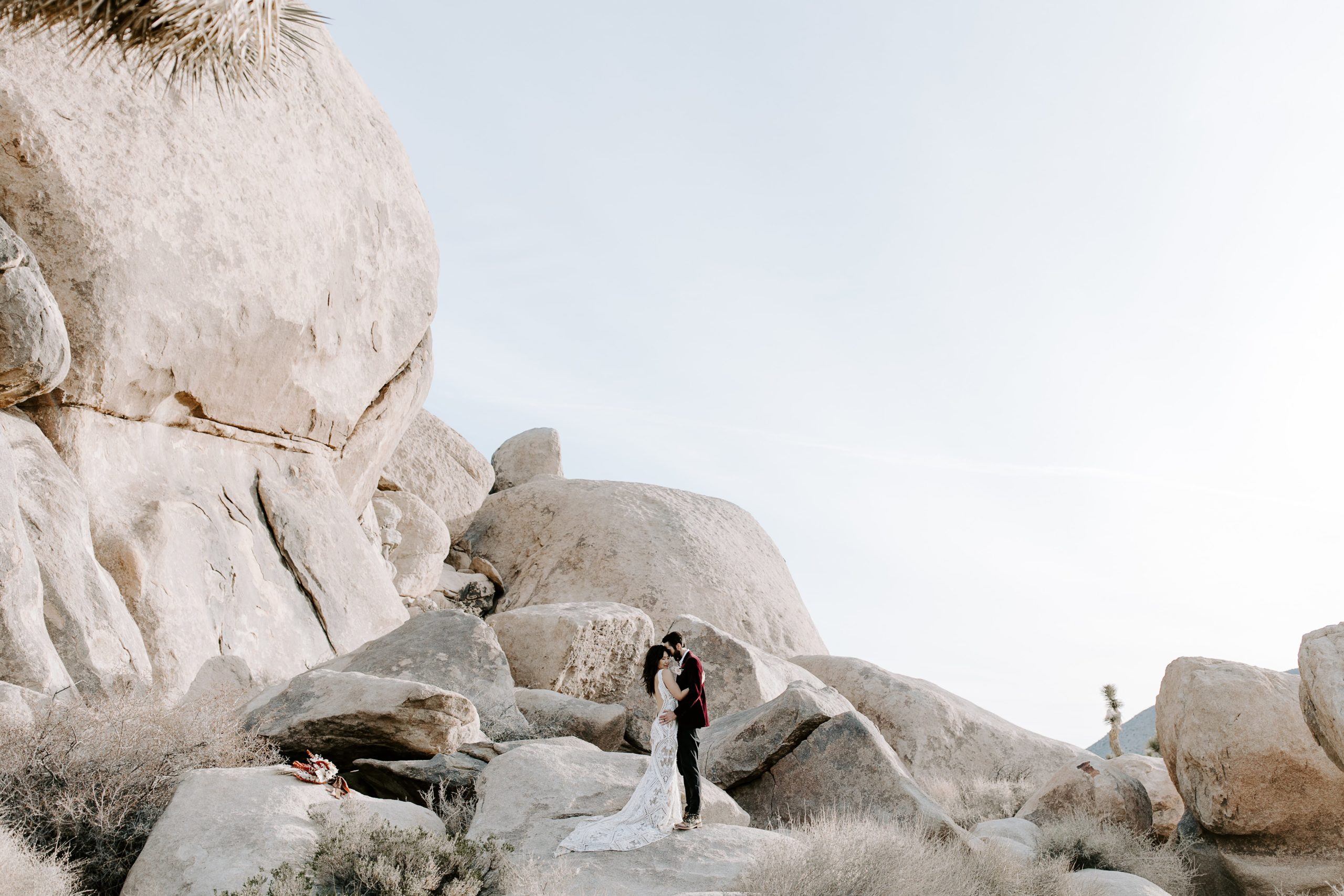 Man and woman embracing in a hug as they stand atop a large neutral colored rock during their hiking elopement in Joshua Tree National Park in California