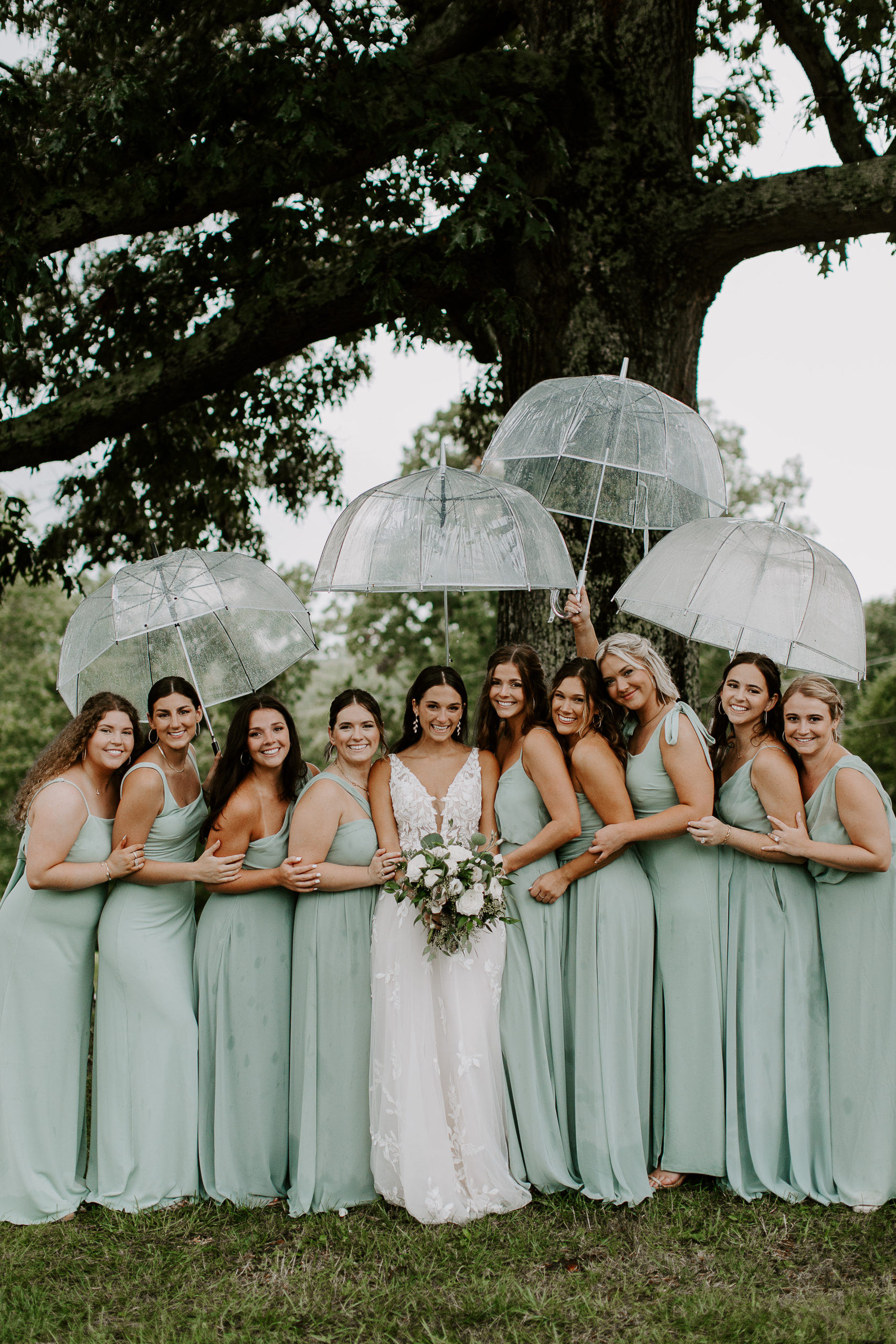 Woman standing in the middle of her bridesmaids as they all lean into her and hold clear umbrellas over their heads during a rainy wedding