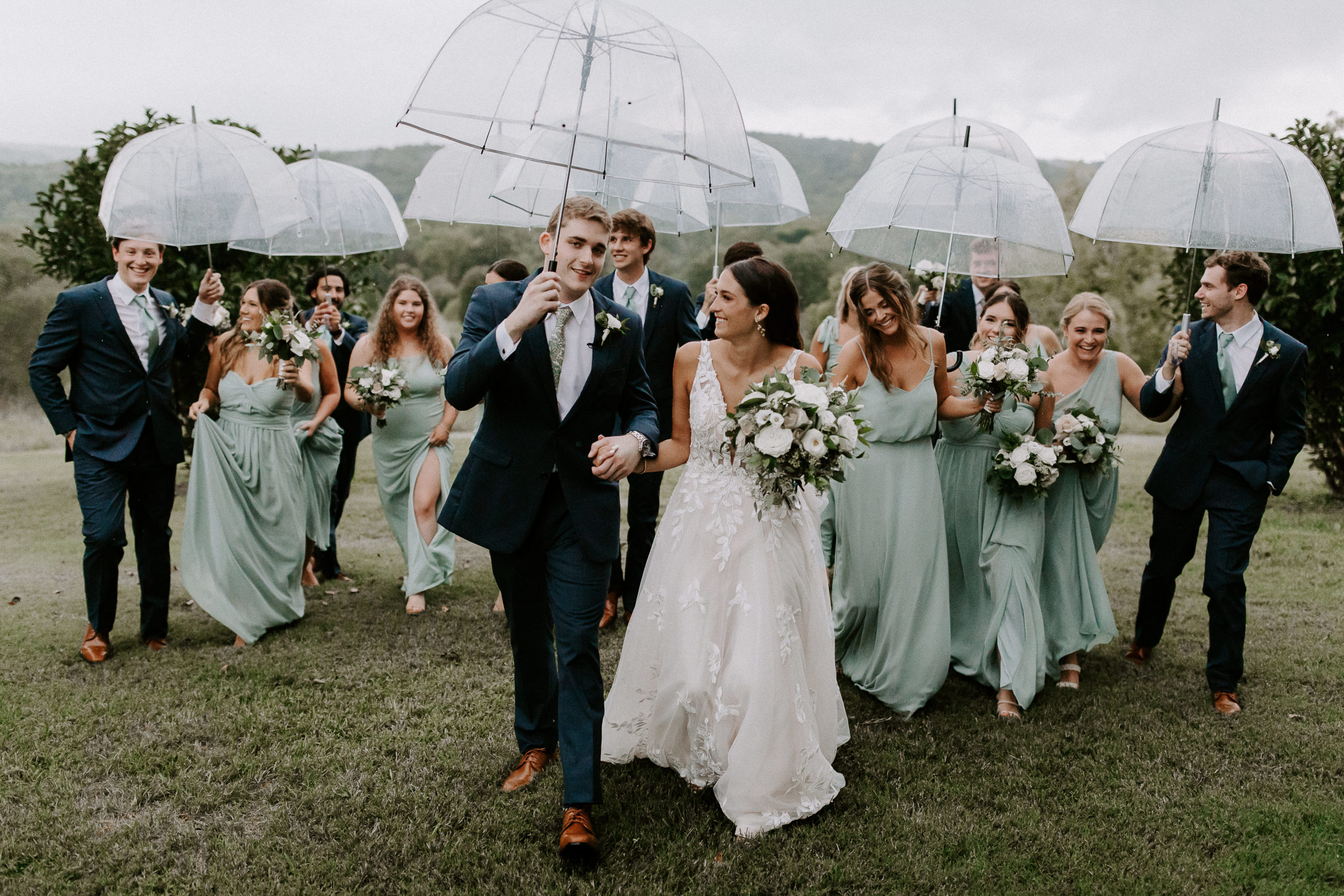 A bride and groom walking through a field followed by their wedding party and everyone is holding umbrellas