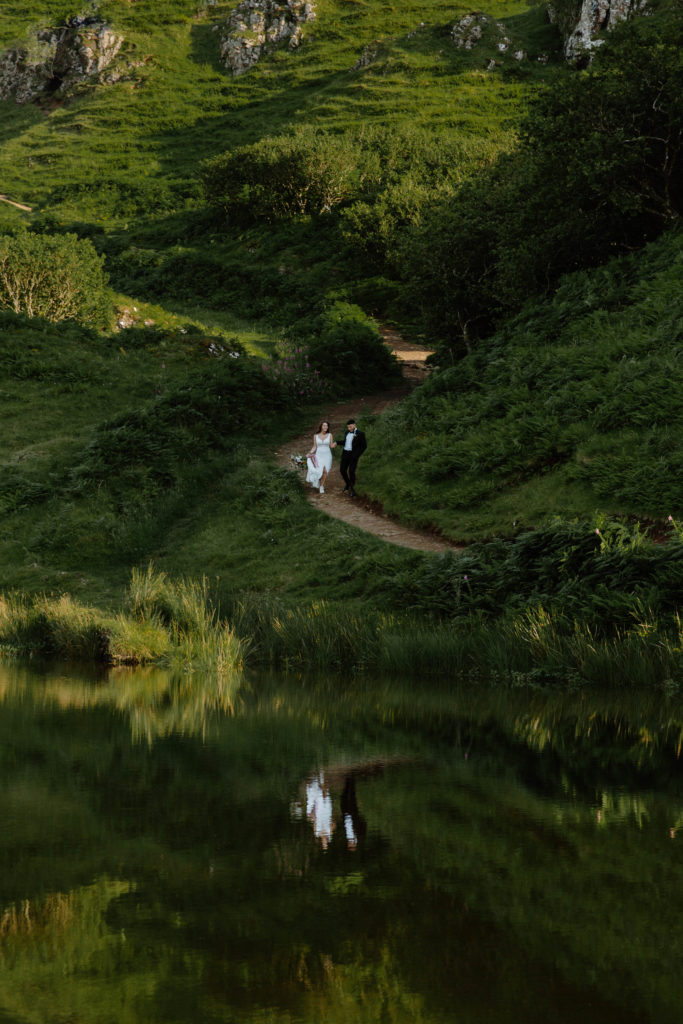 A couple in wedding attire walking down a path with large rock formations behind them and a large body of water in front of them that has their reflection in it during their Scotland elopement