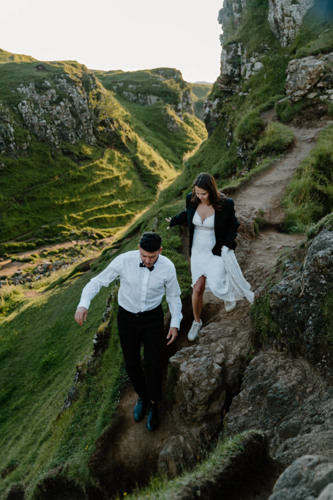 A couple in wedding attire walking down a cliffside embankment at the end of their all day elopement in the Isle of Sky Scotland
