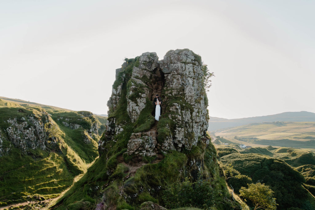 A man and a woman in wedding attire standing at the top of a large rock formation that is jutting out against the Scottish landscape during day 1 of their adventure elopement in Scotland