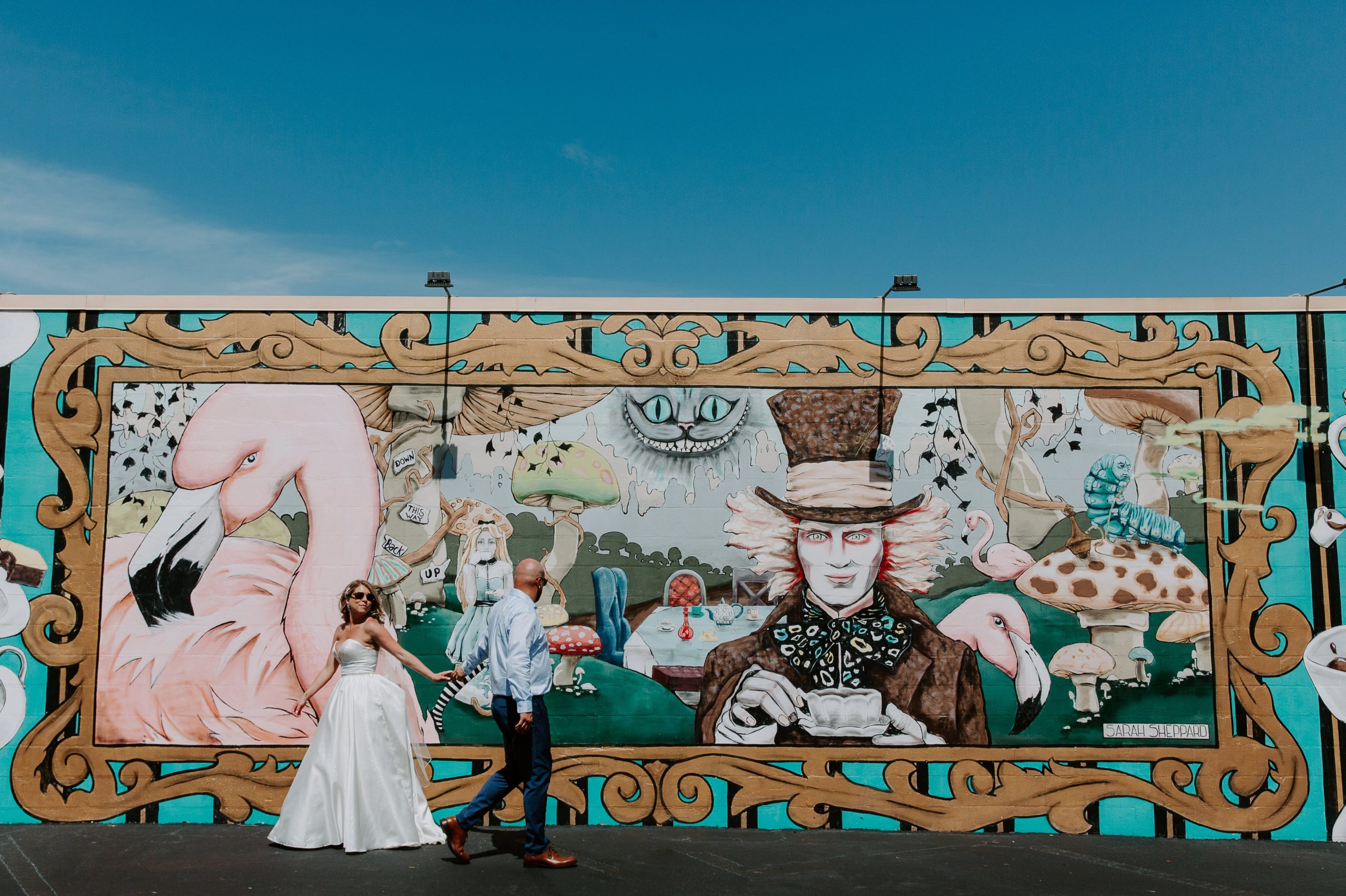 Couple walking past each other and reaching out for the other standing in front of a Mad Hatter mural in downtown St. Petersburg