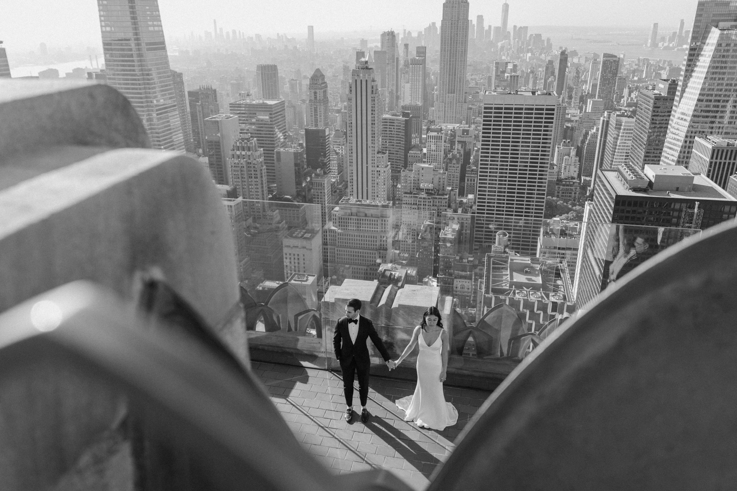 Newly wed couple standing hand in hand on the Top of the Rock platform in NYC with the entire cityscape in the background during their New York wedding