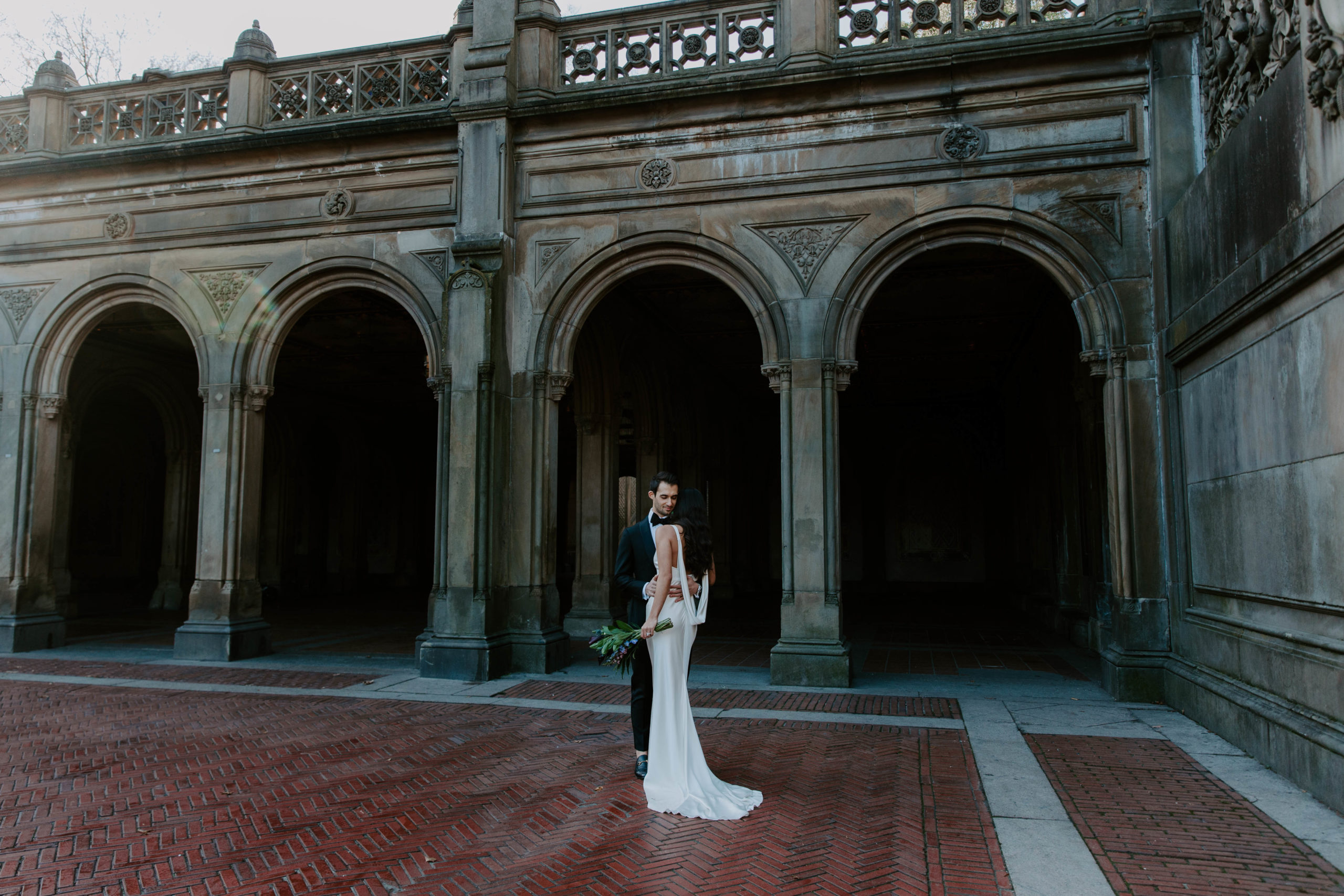 Newly wed couple curled up to each other standing in front of Bethesda Terrace in Central Park during their elopement