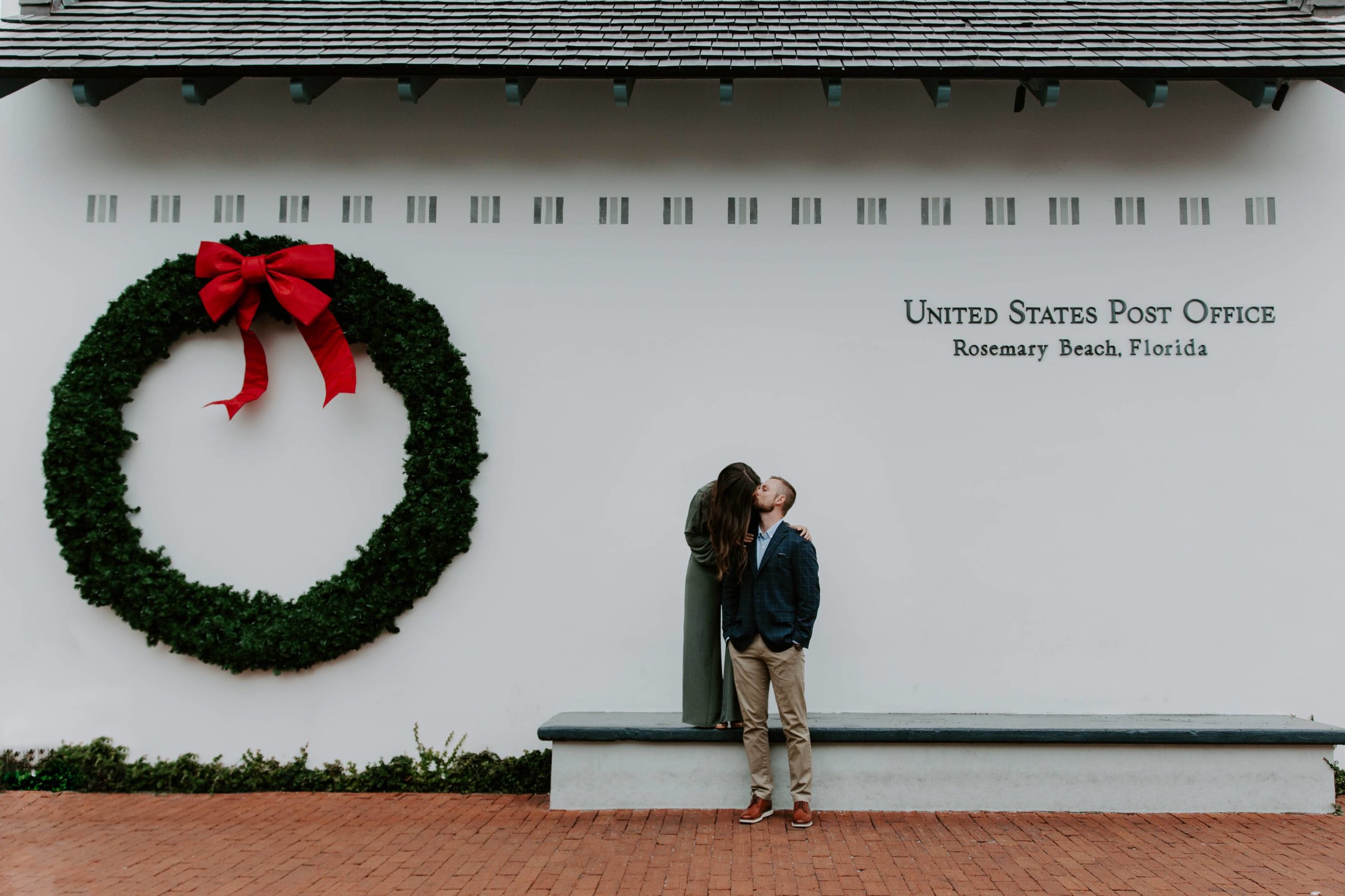 Couple standing in front of the Rosemary Beach Florida post office that has a big wreath on it the woman is standing up on a bench in the man standing in front and the woman is leaning down to give her partner a kiss during their Rosemary Beach engagement photos