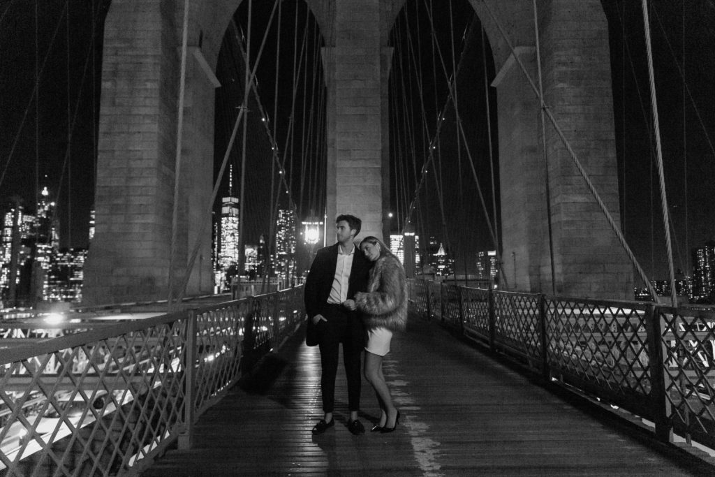 A couple standing in the middle of the Brooklyn bridge with a big arches in the New York City skyline behind them with a woman hugging onto her partner's arm and hammer looking off into the distance during their nighttime photo shoot