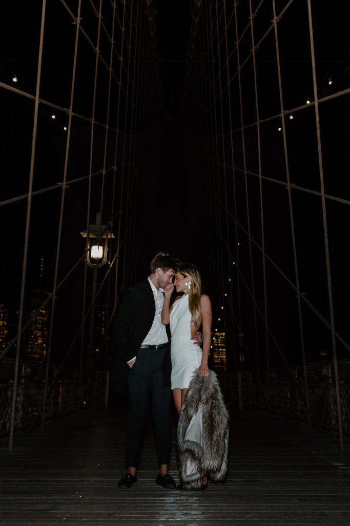 A man with his arms wrapped around his partners waist as she is leaning into him and holding her hand to his face and then her other hand she is holding her fur jacket as they are surrounded by the New York City lights as they stand on the Brooklyn Bridge during the couples photo shoot