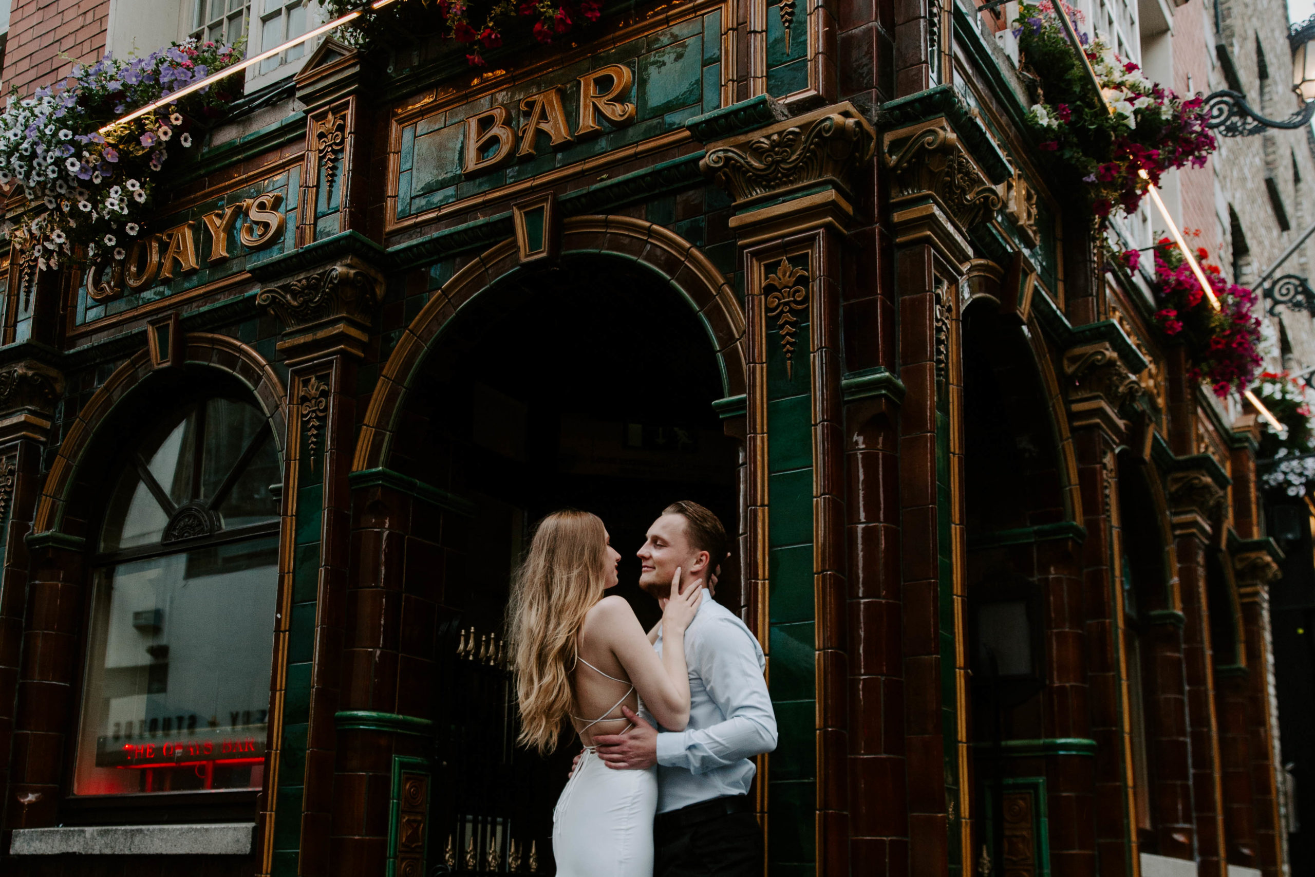 A woman in a wedding dress leaning up against her partner with her hand resting on his face as he looks at her lovingly and they are both leaning up against a pillar of a bar in downtown Dublin during their urban wedding in Ireland