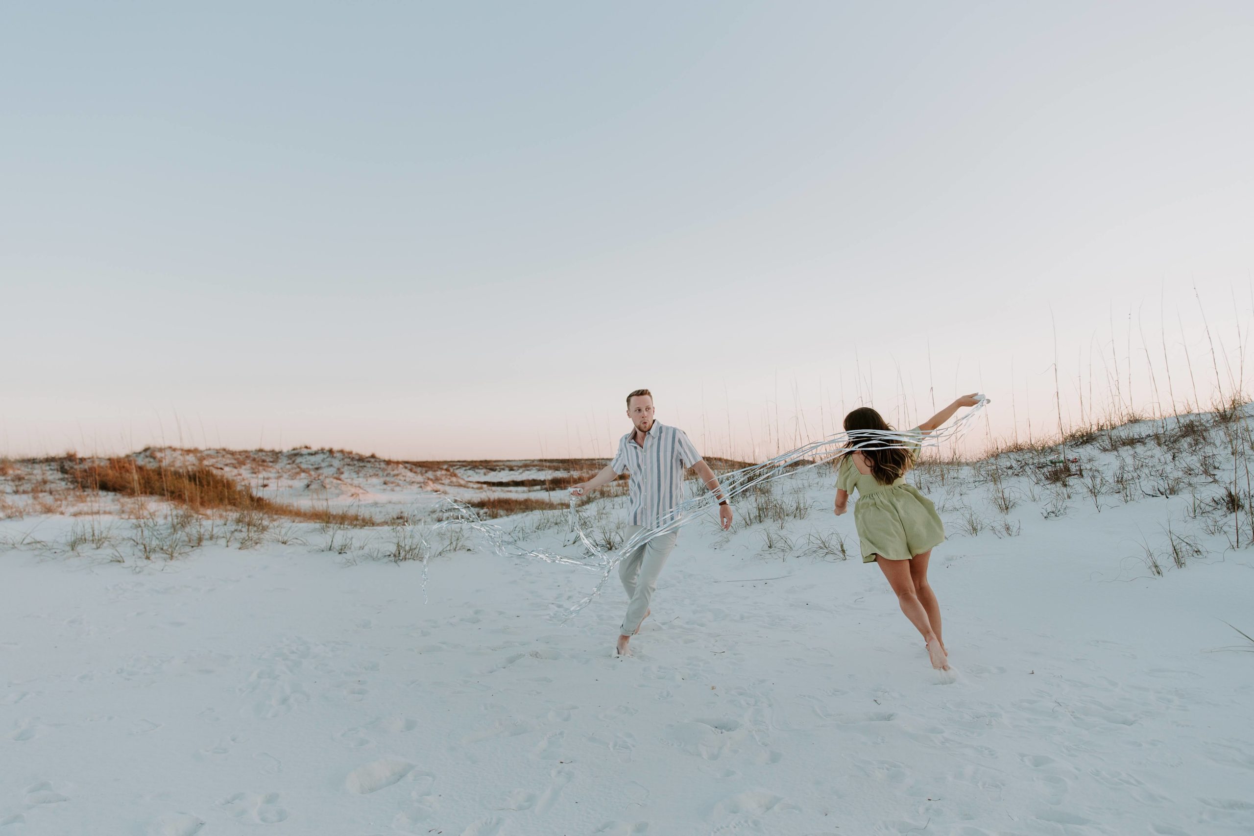 A newly engaged couple running around the beach making silly faces and holding streamers as they celebrate getting engaged during their destination engagement in Florida