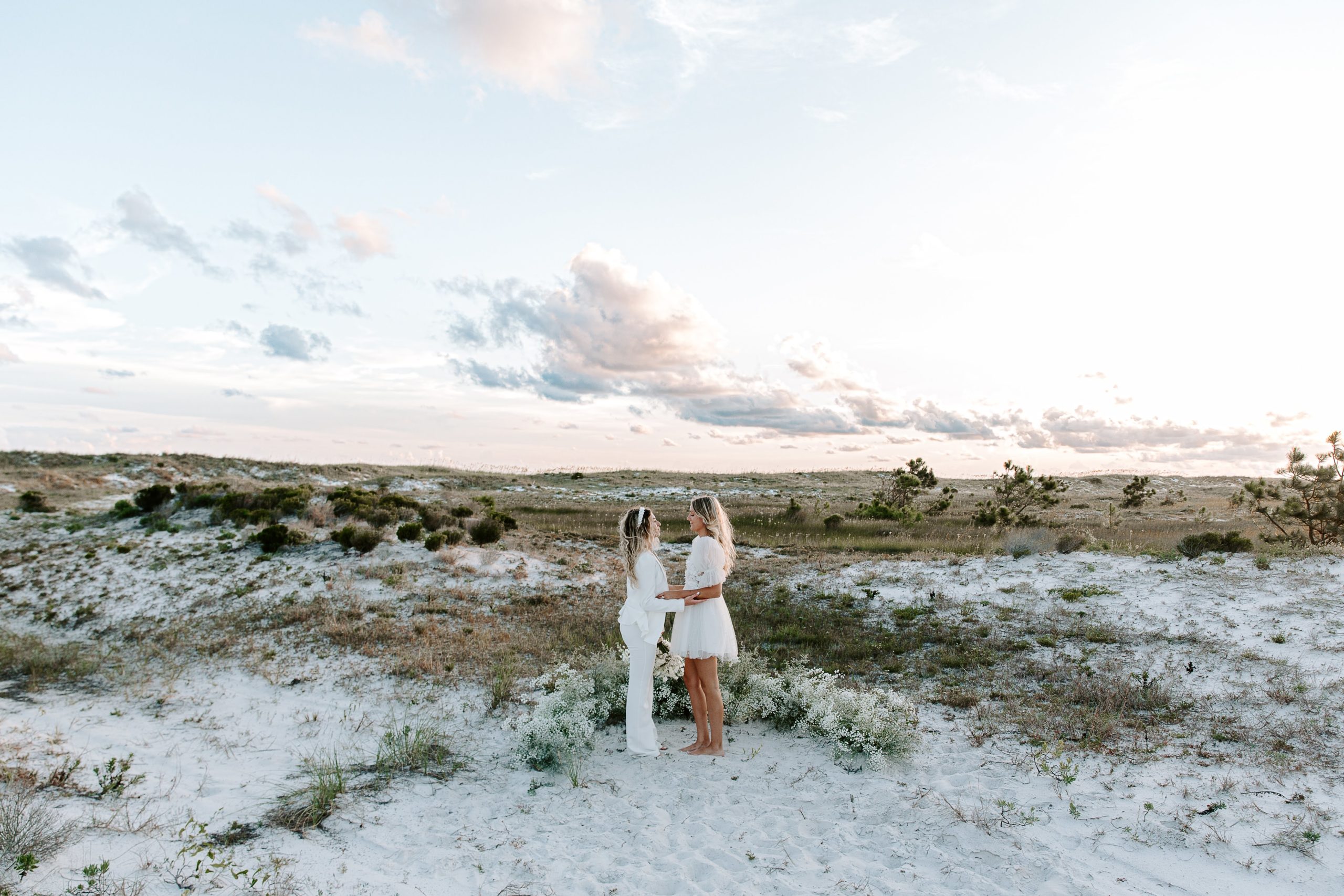 A couple standing in their wedding attire with their arms intertwined as they say their vows in the sun goes down behind them during their beach elopement in the Florida panhandle