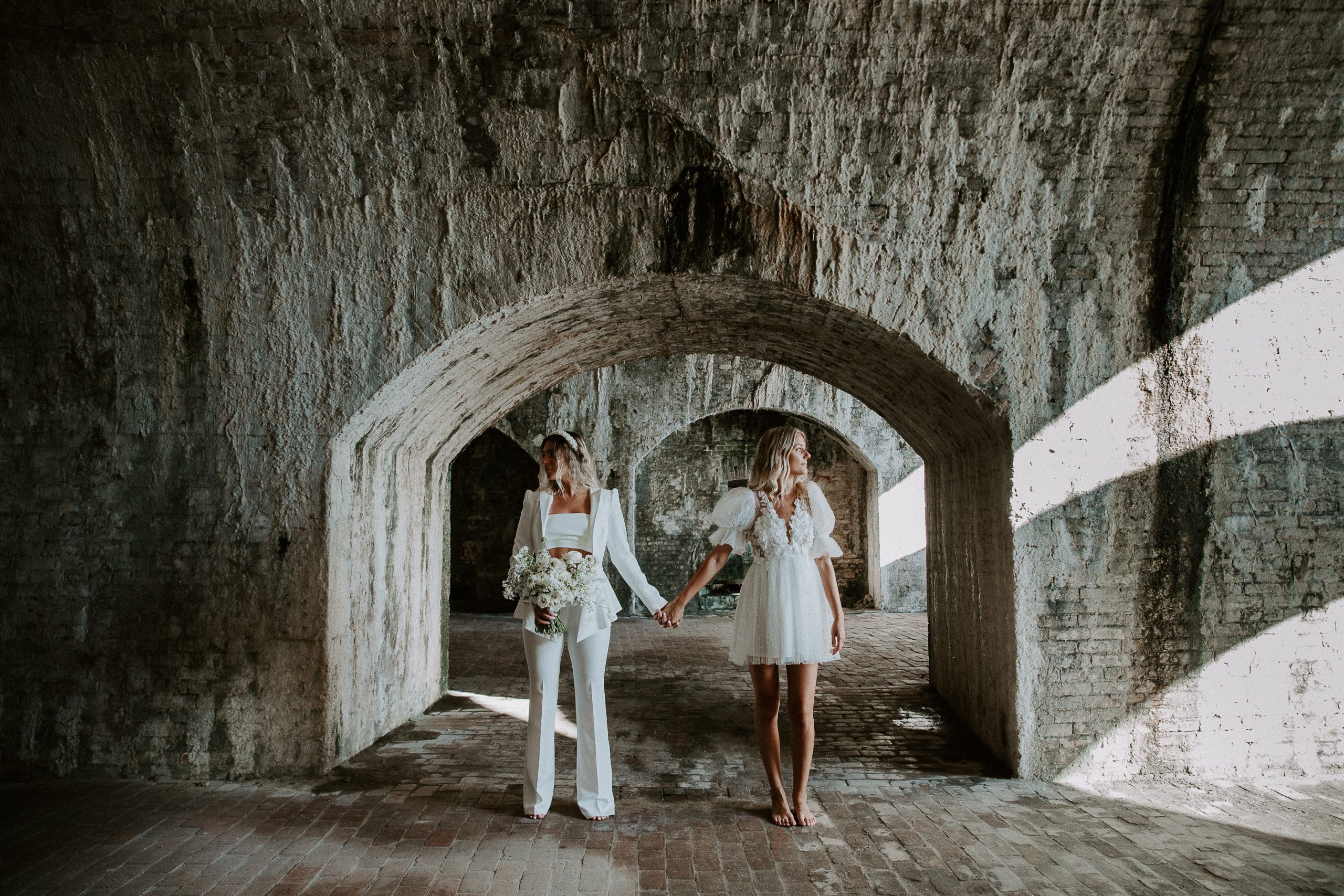 Couple standing hand-in-hand looking opposite directions of each other both in wedding attire standing under an arch in Fort Pickens during their Florida elopement