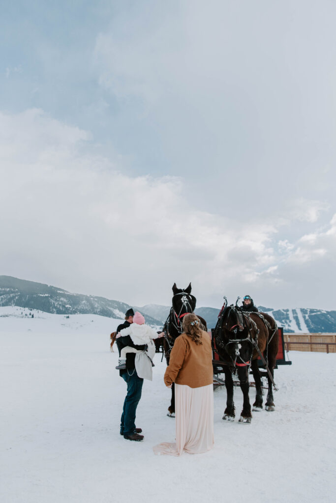 A couple standing holding their daughter as a pet horses after a carriage ride through the elk refuge in Jackson hole during their destination Wyoming elopement