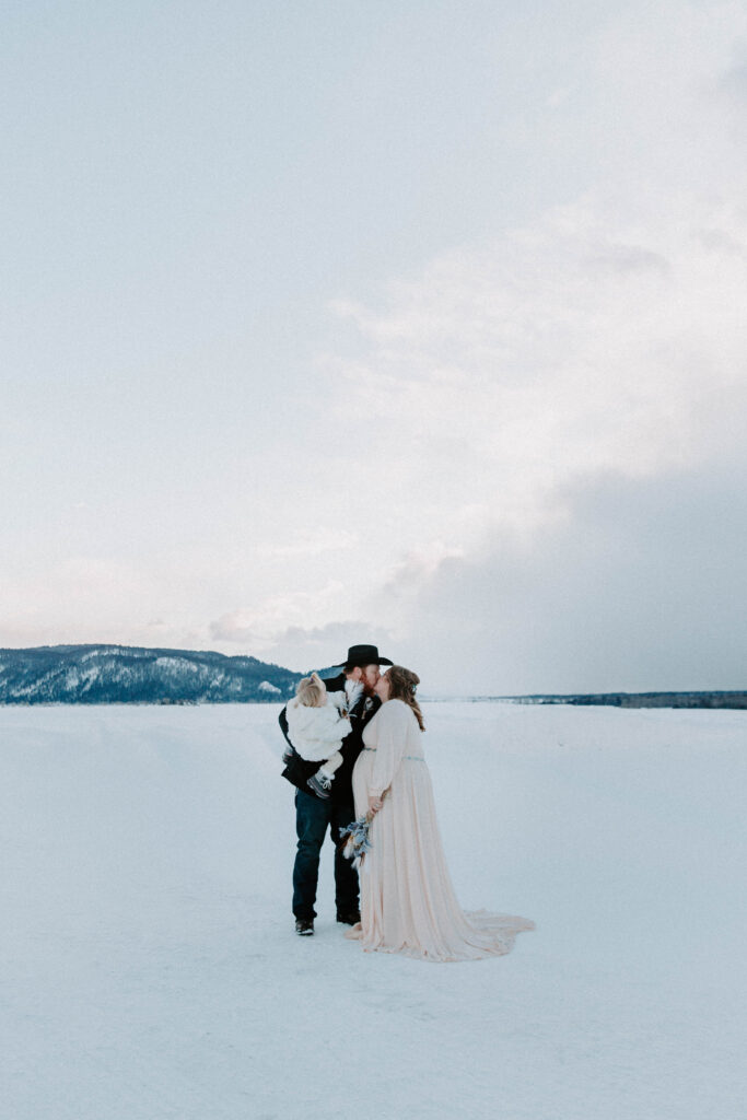A couple of sharing a kiss as they stand on the snow and hold her daughter after they said their vows to each other with the mountains in the background during their national Park elopement in Wyoming