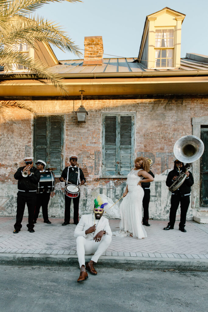 A man sitting down on the curb holding a masquerade mask over his face as his bride is standing behind him and the second line is playing all around them as the sun sets on their French Quarter wedding 
