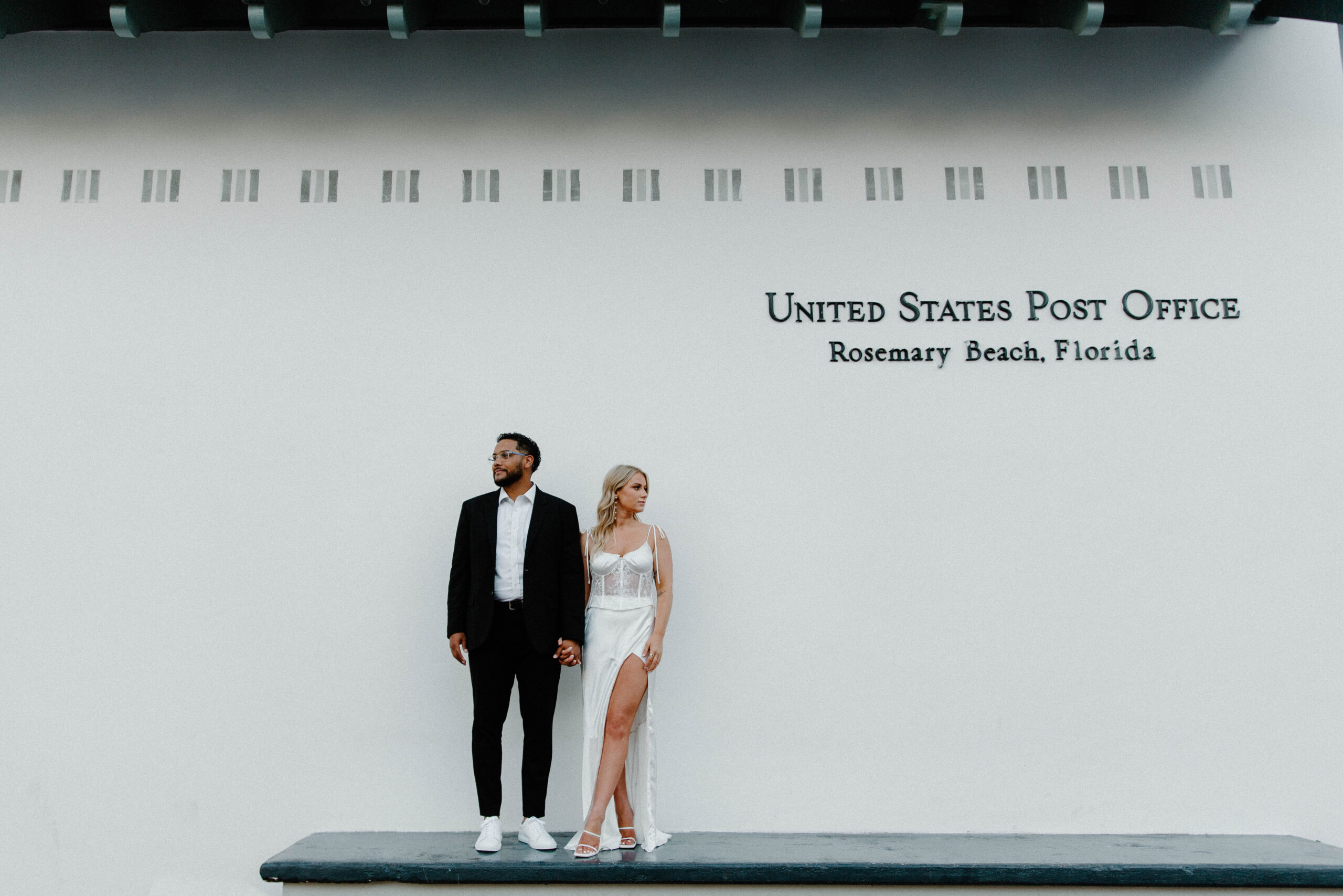A newly engaged couple standing on the Rosemary Beach post office ledge holding hands and looking opposite directions of each other with a man in a black suit and the woman in a white dress