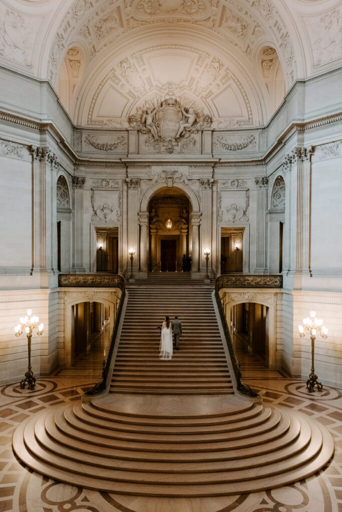 A couple in wedding attire walking up the large staircase in the center of San Francisco City Hall during their weekday morning elopement in California
