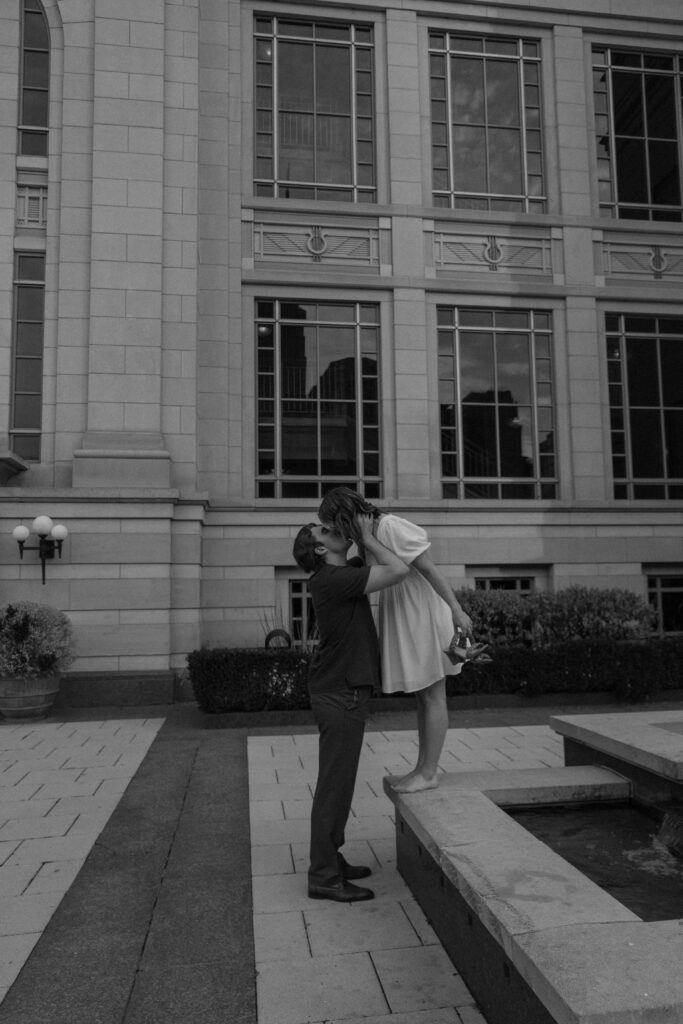 Woman standing on the edge of a fountain with her partner standing on the ground in front of her as she bends down to give him a kiss during their downtown Nashville engagement photos in Tennessee