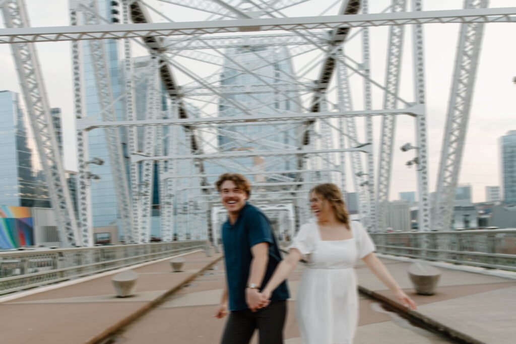 Couple holding hands and running down the Pedestrian Bridge in Nashville as they are laughing during their Tennessee engagement photos