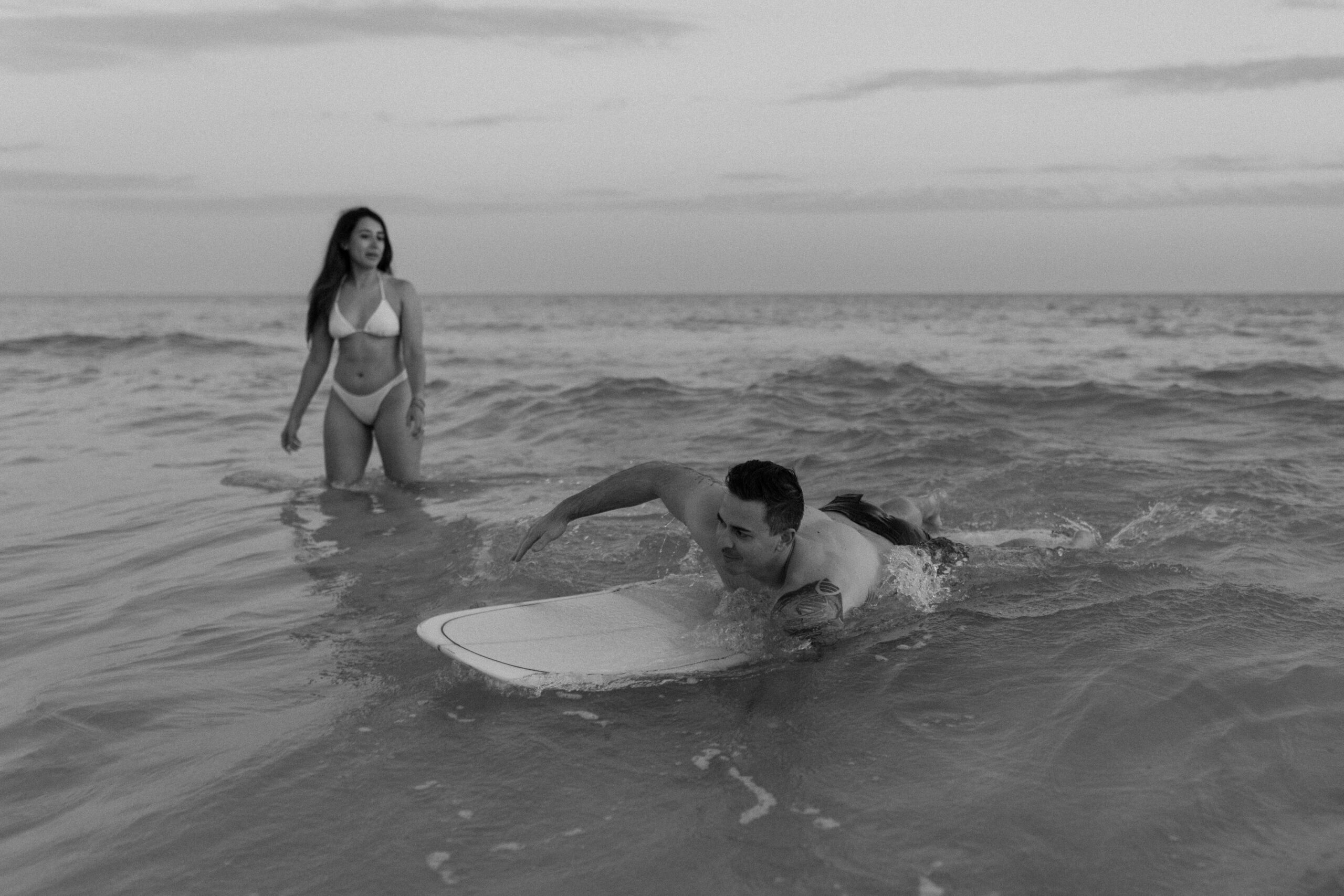 A man riding waves on a surfboard as his partner is next to him in the water during their sunset couple photoshoot on the beach