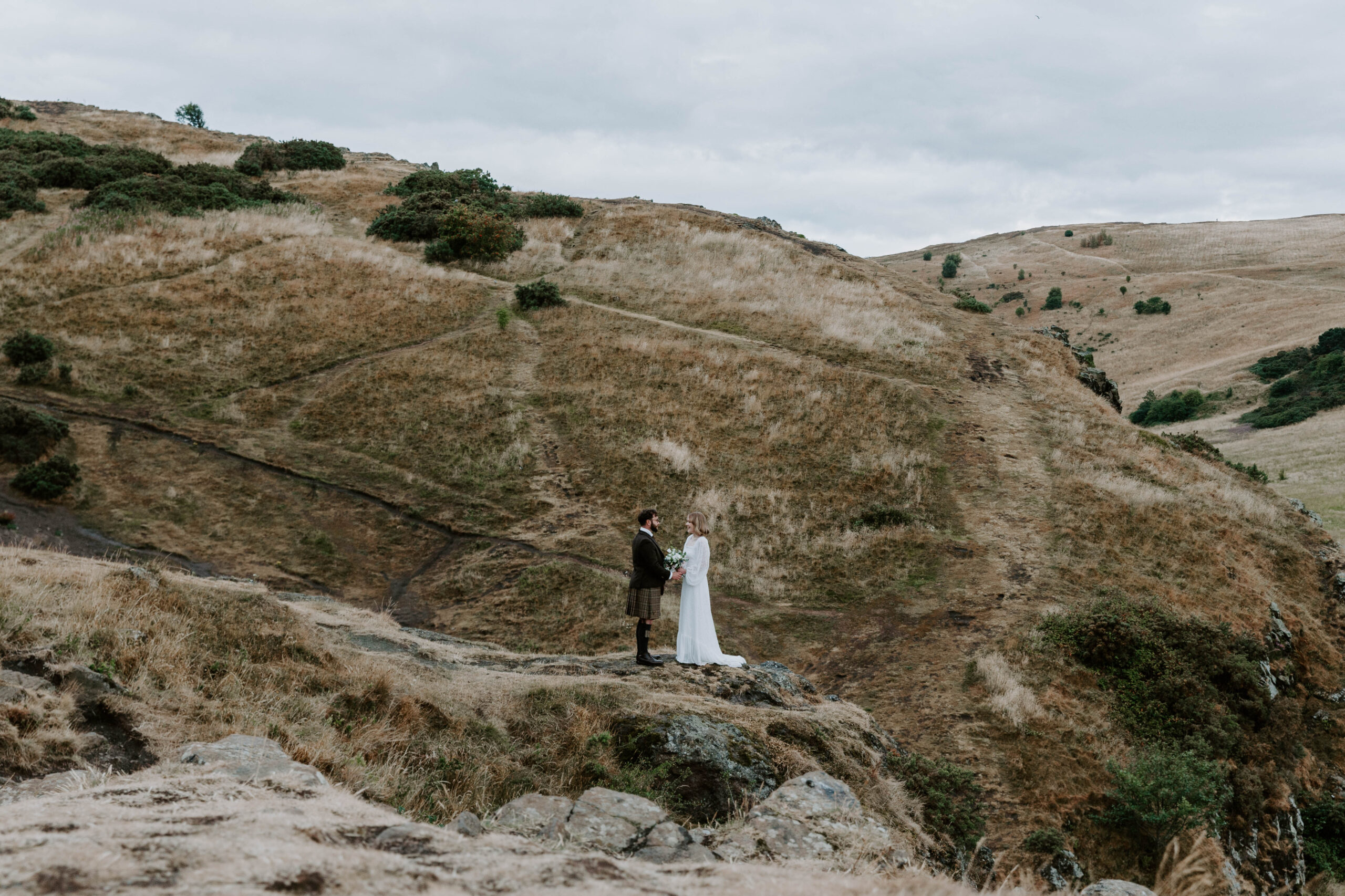 A couple holding hands and standing on a ledge overlooking Edinburgh during their Scotland elopement