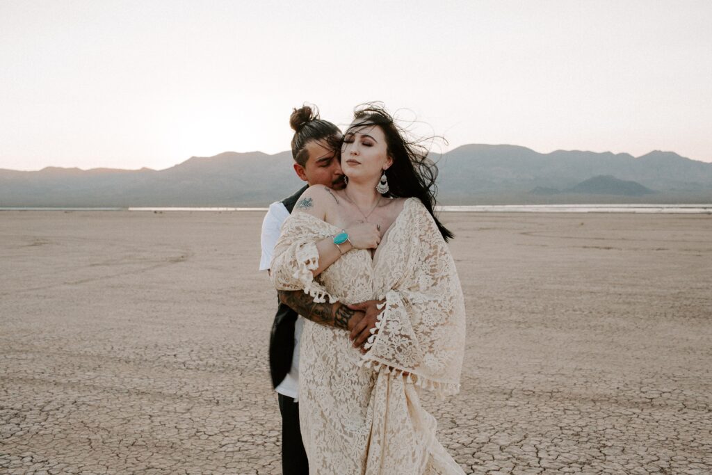 A man with his arms wrapped around his partner as the wind whips her hair and wedding dress around in the desert of Nevada during their Las Vegas elopement