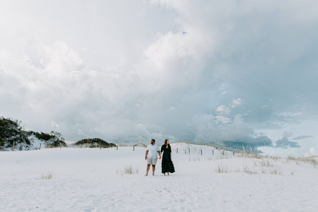 A couple standing hand in hand as storm clouds gather in the background during their morning beach couple session