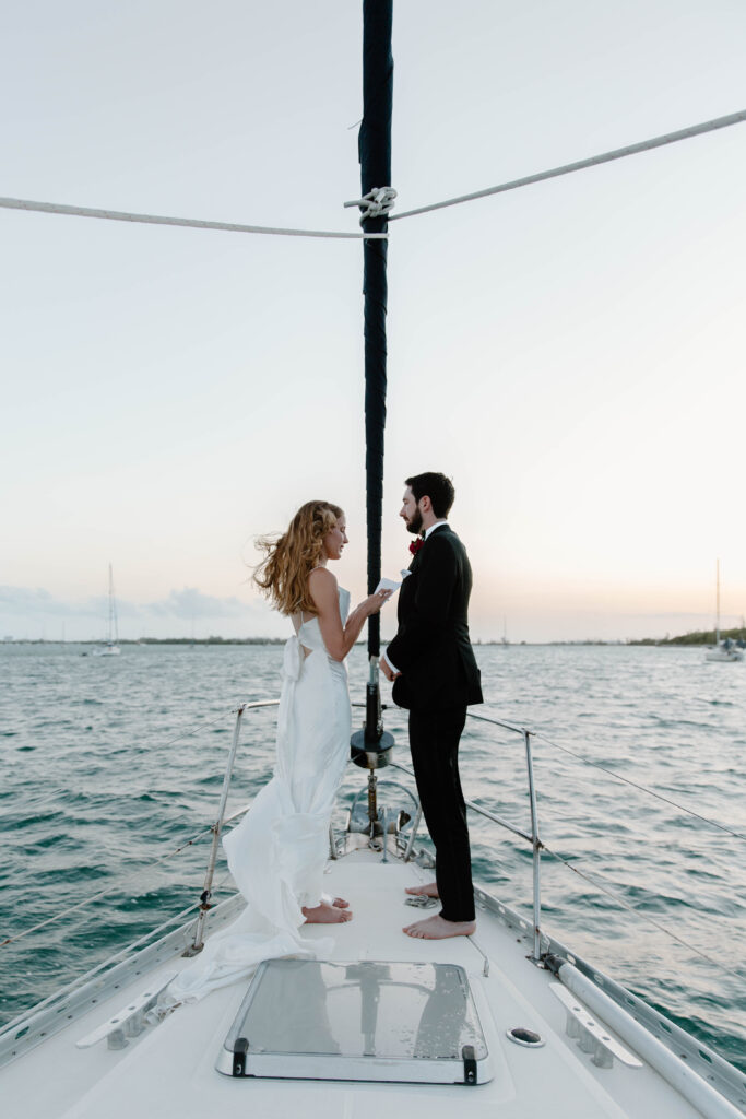 A couple standing at the front of a sailboat as the sun rises reading vows to each other during their Key West adventure elopement