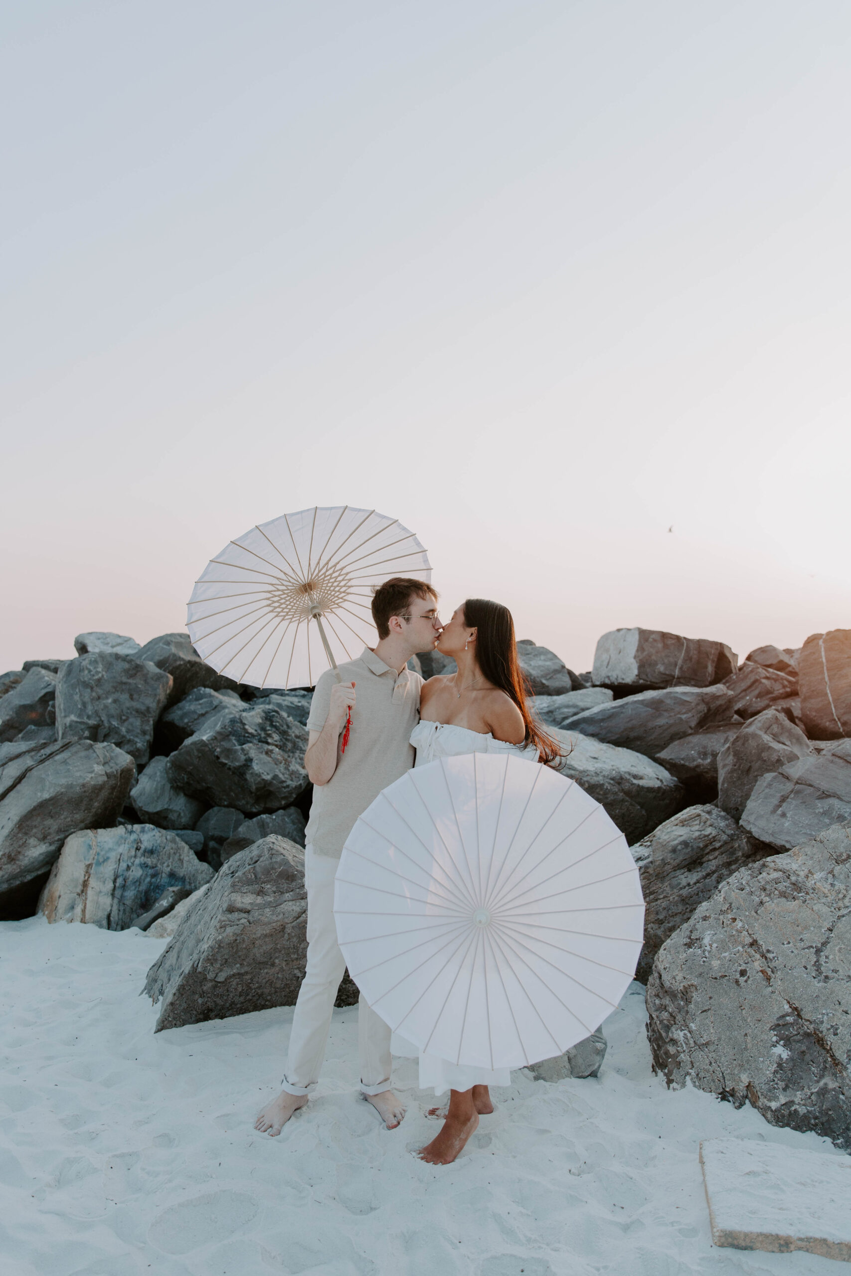 A couple holding parasols and sharing a kiss as the sun dips below the horizon during their Destin Florida couple photo shoot