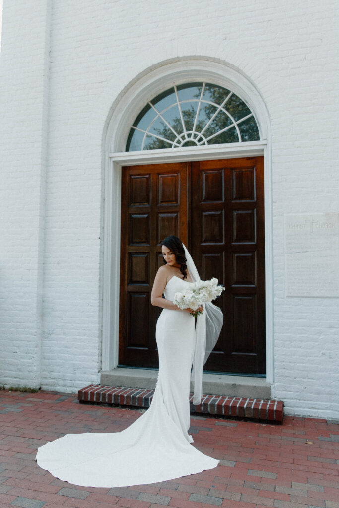 A bride standing in front of large wooden doors of old Christ Church looking down at the ground as she holds her white bouquet during her destination wedding in Florida