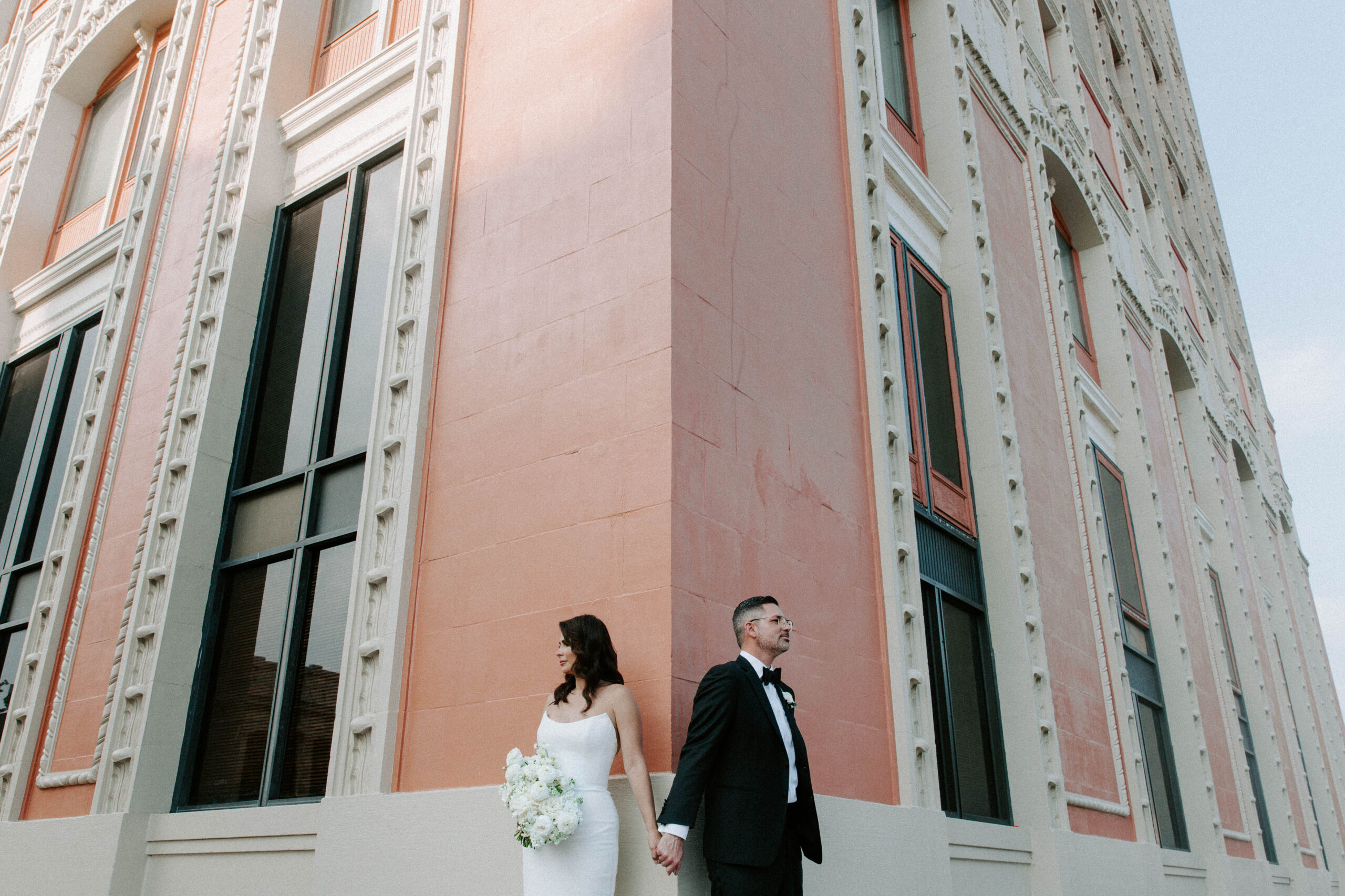 A couple standing on different sides of a wall but holding hands around the corner during their downtown Pensacola wedding portraits