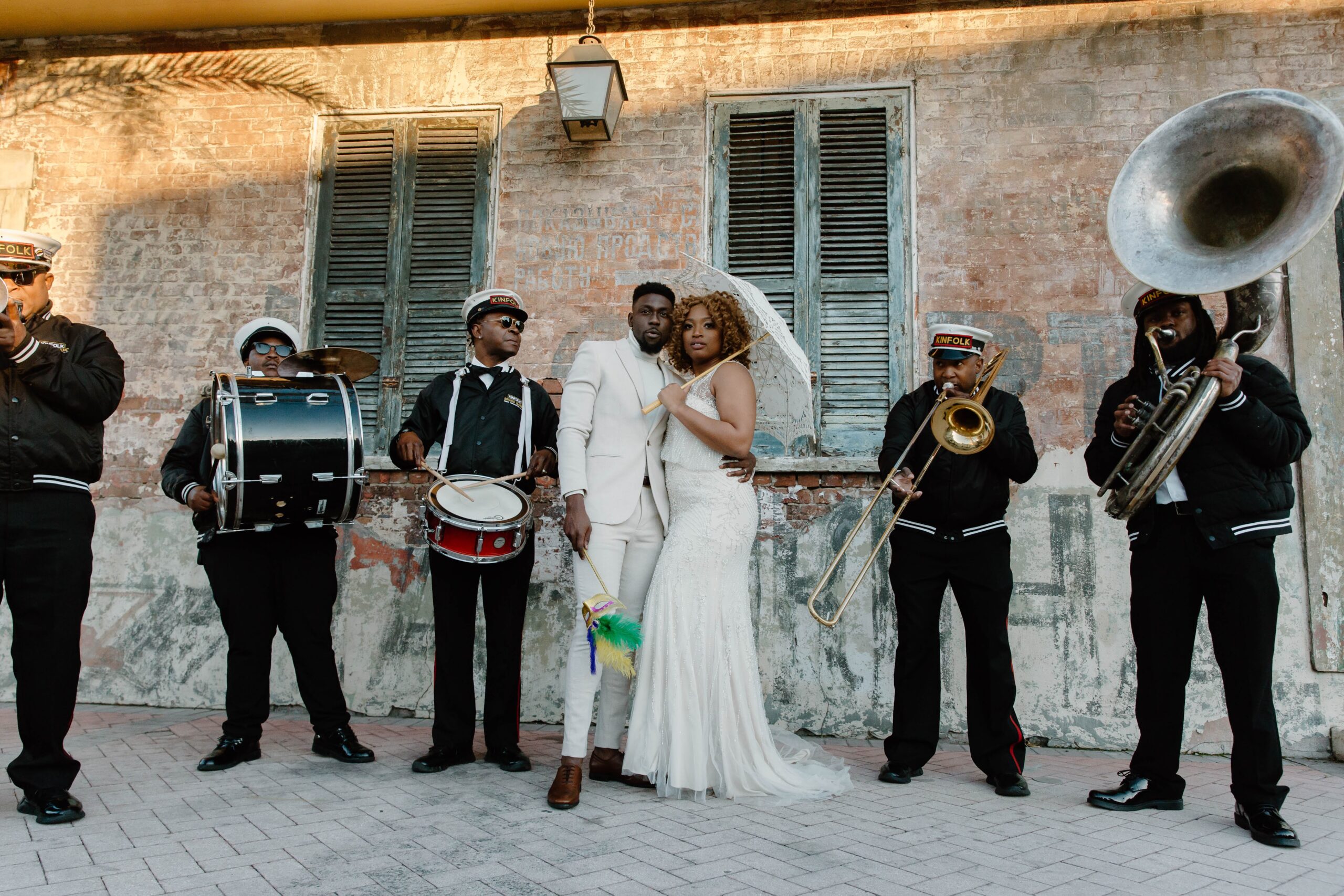 A couple standing close together and wedding attire as they are surrounded by a brass band, and the woman is holding a parasol above her head, and the man is holding a Mardi Gras mask during their urban New Orleans elopement