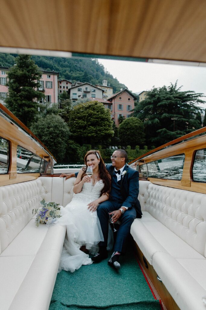 A couple sitting in a boat with a man's arm placed behind his wife and she is holding a wine glass as they take a boat ride on Lake Como during their Italy intimate wedding