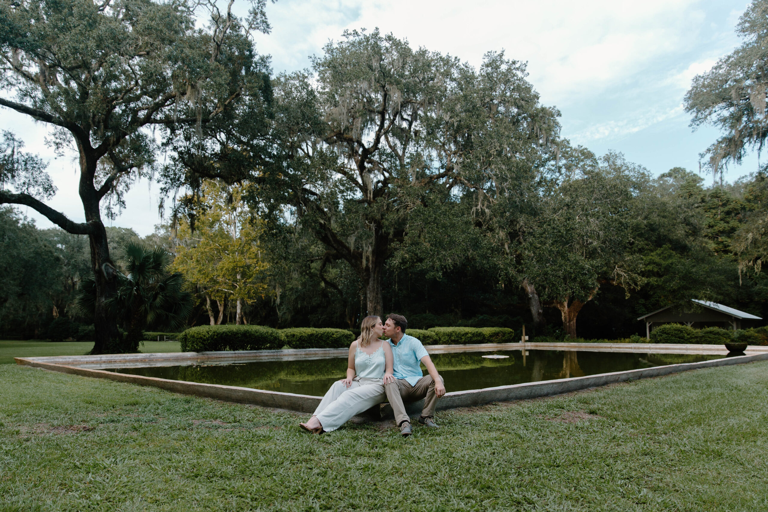 A couple of sharing a kiss as they sit on the edge of a large fountain surrounded by large oak trees during their Florida engagement photos